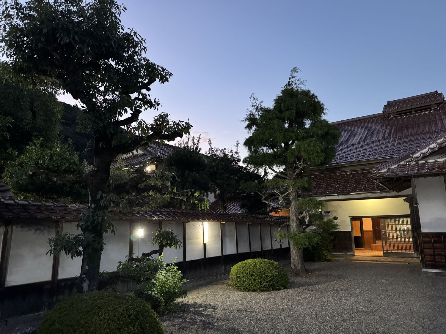 白壁の土塀に沿って灯りがともり、奥の玄関から明かりが漏れる夕暮れの屋敷